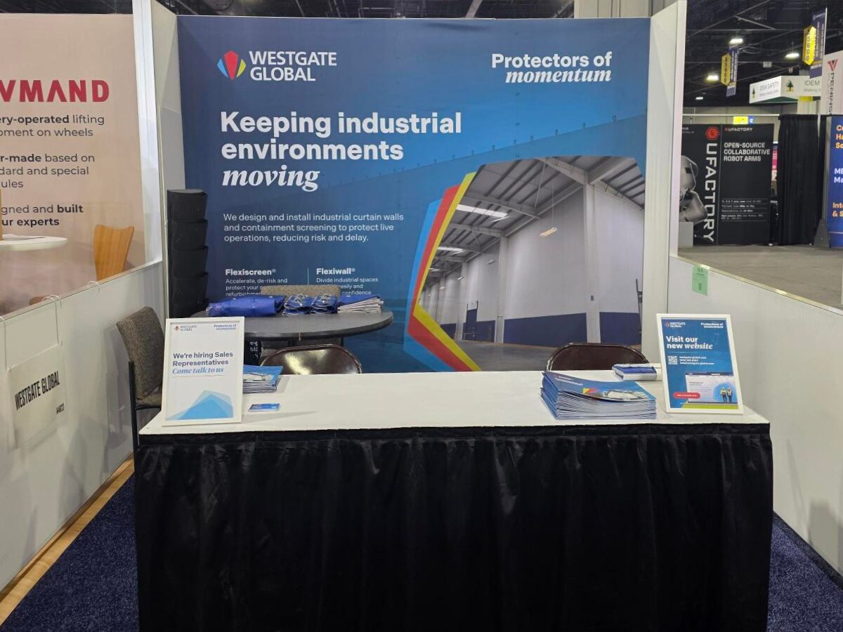 Westgate Global exhibition stand at the MODEX trade show. A large blue backdrop displays the company logo, tagline "Protectors of momentum," and the headline "Keeping industrial environments moving." The stand features a table skirted in black with brochures and promotional signage. Neighbouring stands are partially visible in the background.