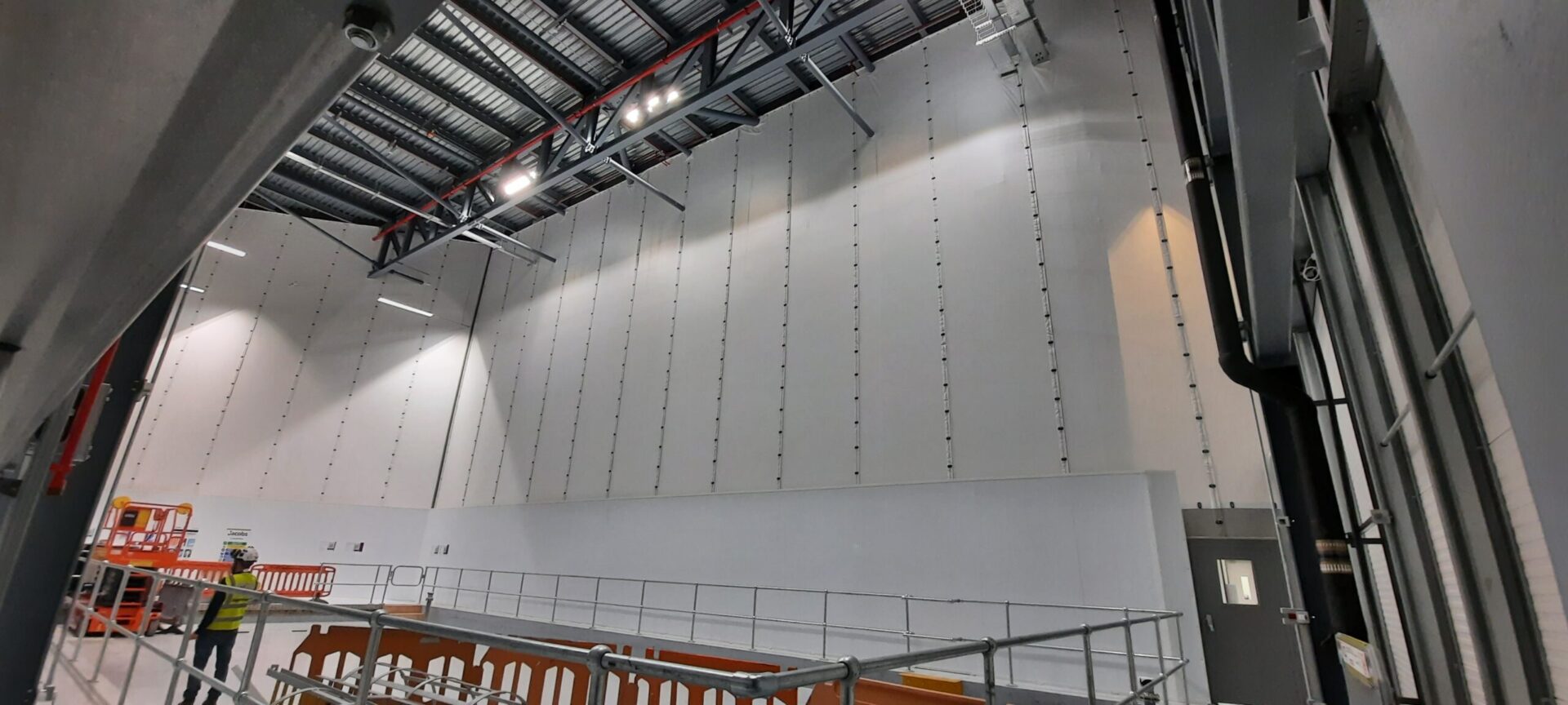 Large-scale Flexiscreen industrial curtain wall system in a pharmaceutical manufacturing facility, showing floor-to-ceiling white PVC partition panels suspended from steel roof trusses with vertical track guides, creating a clean-room grade dividing wall. A worker in high-visibility PPE and a safety helmet is visible in the foreground alongside temporary scaffolding barriers and an orange scissor lift platform, with industrial LED lighting illuminating the high-bay interior.