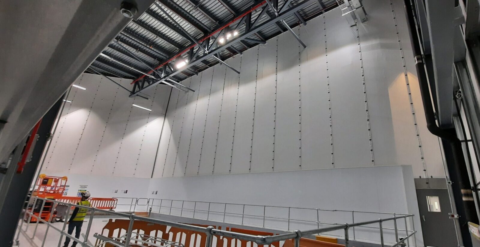 Large-scale Flexiscreen industrial curtain wall system in a pharmaceutical manufacturing facility, showing floor-to-ceiling white PVC partition panels suspended from steel roof trusses with vertical track guides, creating a clean-room grade dividing wall. A worker in high-visibility PPE and a safety helmet is visible in the foreground alongside temporary scaffolding barriers and an orange scissor lift platform, with industrial LED lighting illuminating the high-bay interior.